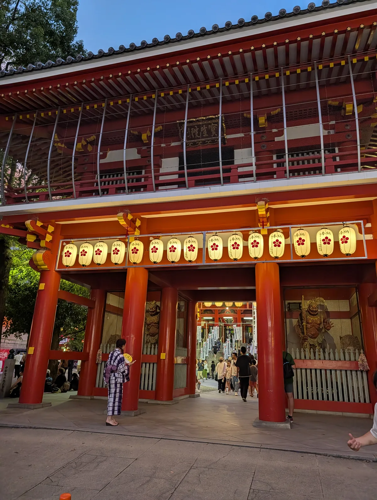 The lantern-lit gate of Osu Kannon temple at dusk during the Osu bon odori, with a visitor in yukata holding a round fan