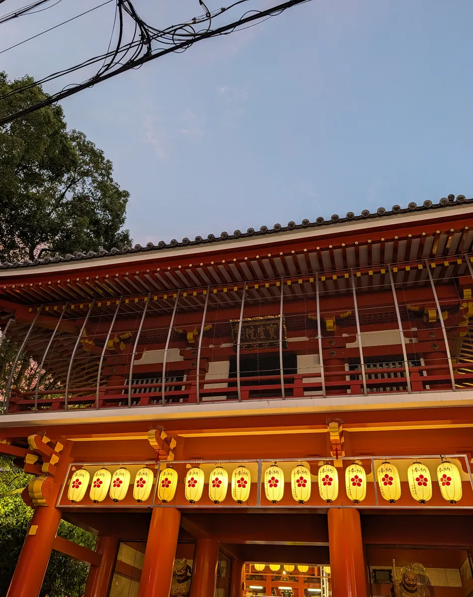 A row of glowing paper votive lanterns marked with a red plum-blossom crest, hung beneath the tiled eaves of the vermilion two-storey gate at Osu Kannon