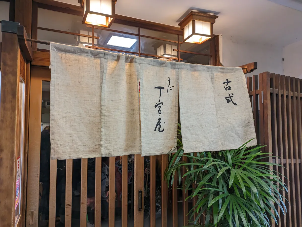 A pale hemp noren curtain brushed with the character for soba hanging across a wooden shopfront in the Osu arcade, with lattice screens, two boxy paper lanterns overhead and a fan palm beside the door