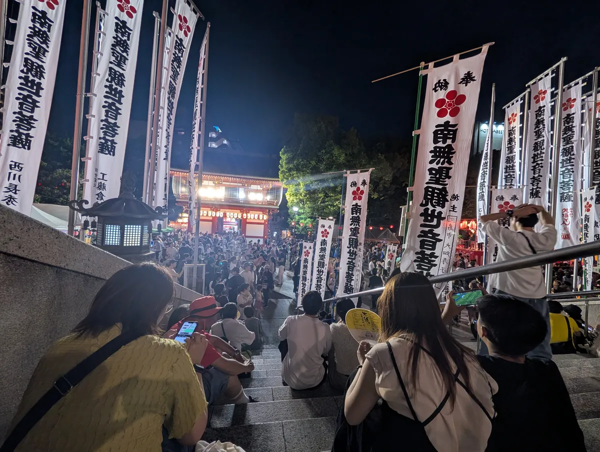 Night crowd seated on the stone steps at Osu Kannon during a summer festival, framed by tall white votive banners, with a yellow paper fan printed for the Osu Summer Festival