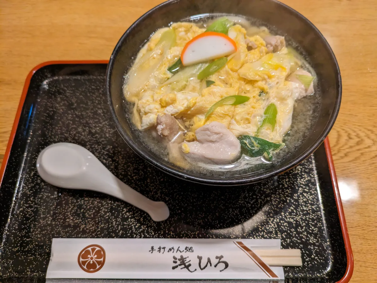 A dark bowl of udon in clear broth with egg dropped through it, pieces of chicken, spring onion and a slice of pink-rimmed fish cake, set on a black tray with a white soup spoon