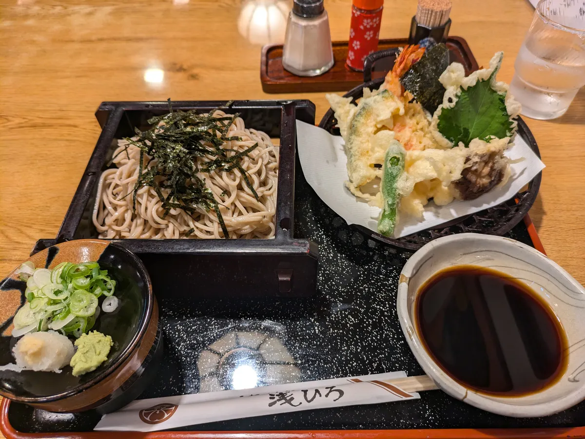 Cold buckwheat noodles topped with shredded nori in a black lacquered box, served with a basket of assorted tempura including prawn, shiso and green pepper, a bowl of dipping broth, grated daikon, wasabi and sliced spring onion