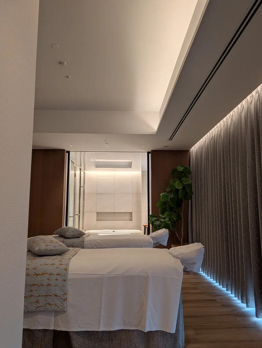 Two side-by-side treatment beds in a couple's room at the TIAD Nagoya spa, seen from the foot of the beds, with a glass-walled bathroom and an oval tub at the far end and a tall leafy plant in the corner