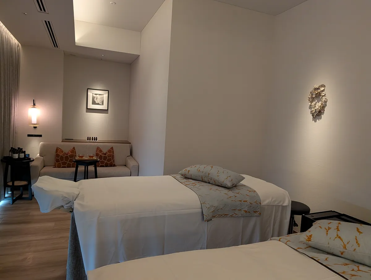 Wide horizontal view of a couple's treatment room at the TIAD Nagoya spa showing two beds, a sofa with tea and a tray of oils in the far corner, a lit framed photograph and a sculptural wall piece