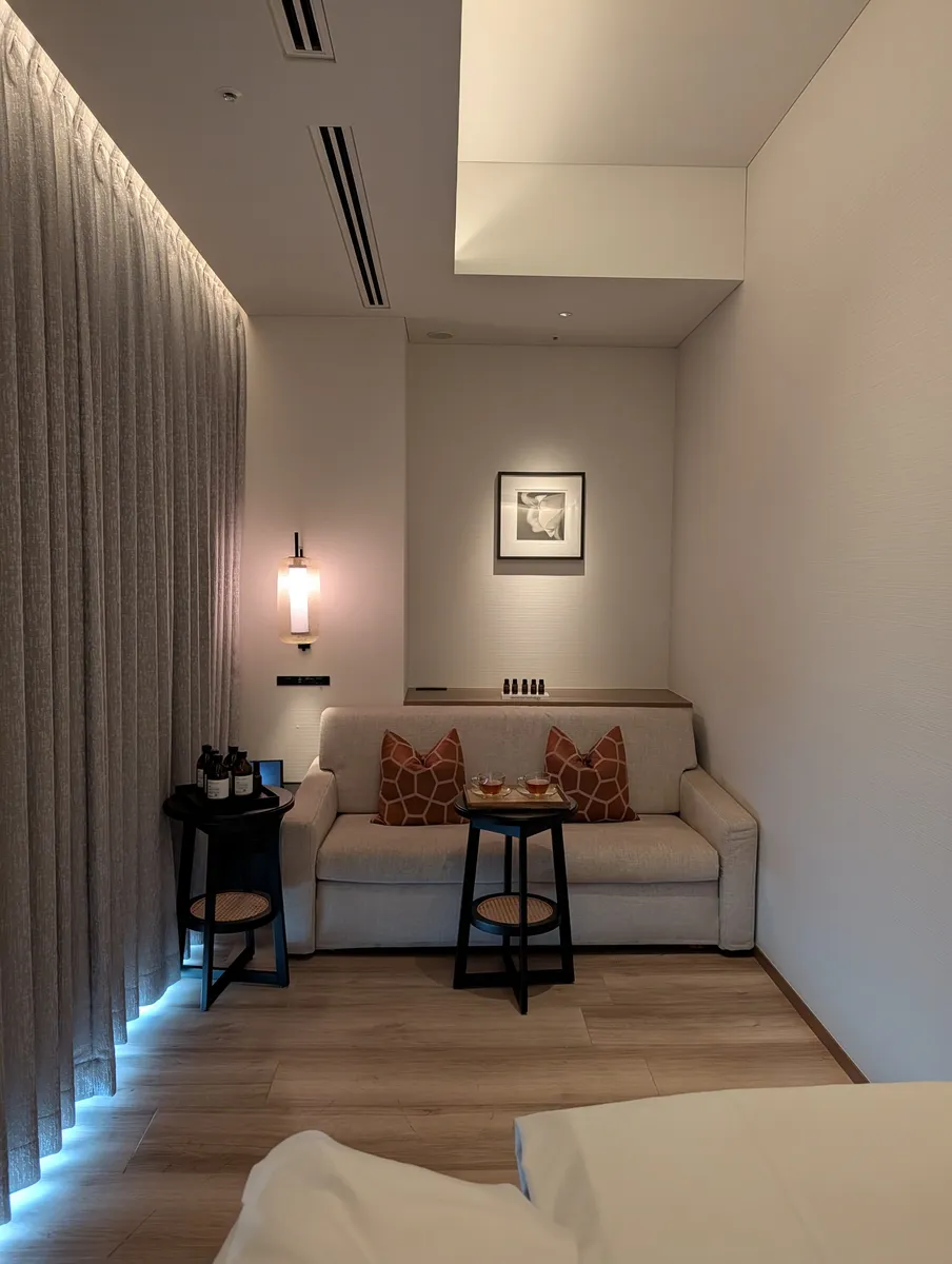 A pale two-seat sofa in a treatment room at the TIAD Nagoya spa, with two small glasses of tea on a wooden tray on a low round table, a stand of oil bottles beside it and a lit framed photograph on the wall above