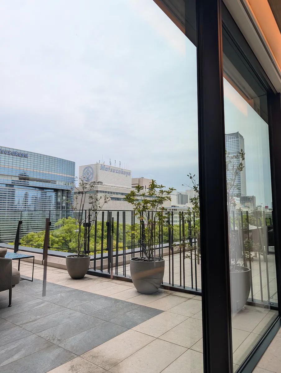 Looking out from inside TIAD Nagoya through a full-height glass opening onto a planted outdoor terrace, with the Matsuzakaya department store buildings and a band of park treetops beyond the railing