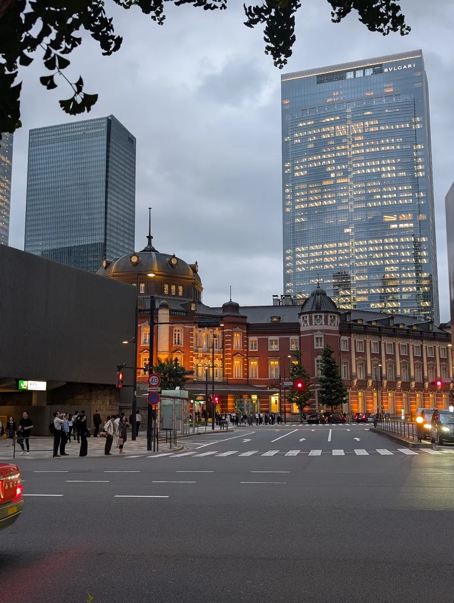 The red-brick Marunouchi front of Tokyo Station lit warm at dusk, with a JR station sign on the near corner and glass towers rising behind it.