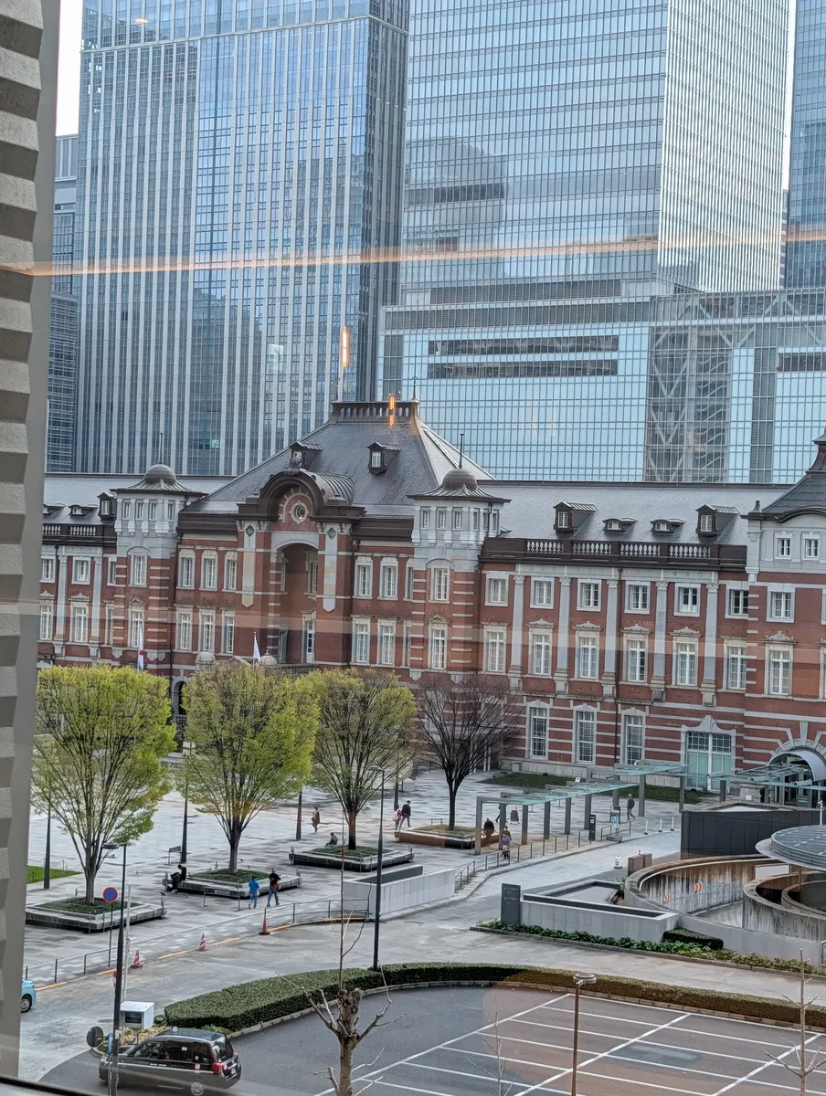Tokyo Station's long red-brick Marunouchi facade and its paved plaza seen from above, framed by office towers, with a few people crossing the square.