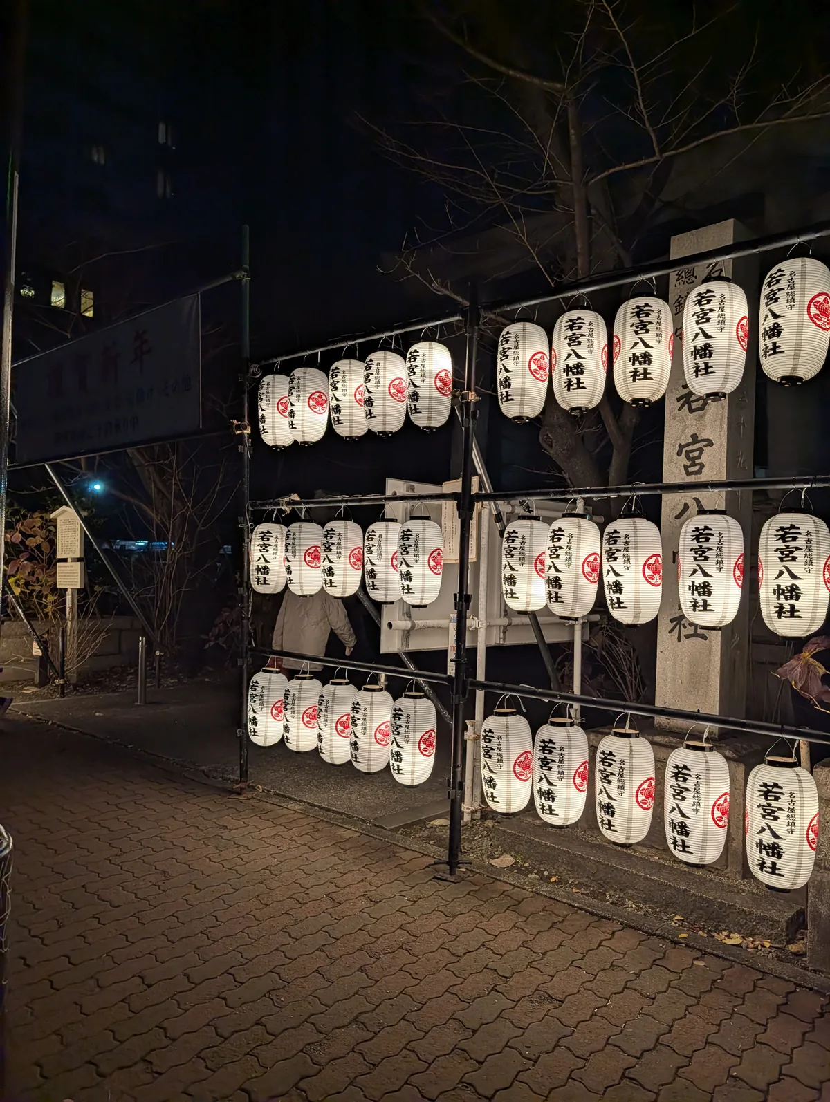 Racks of glowing white shrine lanterns set up at night beside a New Year greeting sign at Wakamiya Hachimansha in Nagoya