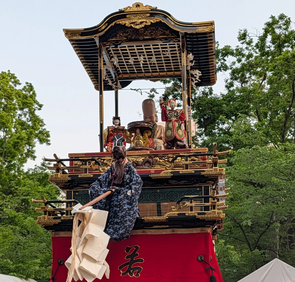 Karakuri mechanical puppets in miniature costume standing on the upper deck of a black-and-gold lacquered festival float, above a red curtain bearing a gold character